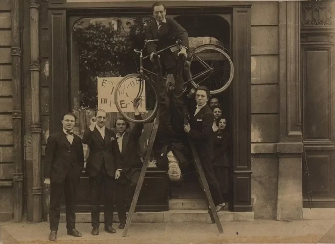 Vernissage de l'exposition de Max Ernst au Sans-Pareil, 1920. De gauche à droite : René Hilsum, Benjamin Péret, Serge Charchoune, Philippe Soupault en haut de l'échelle, Jacques Rigaut (la tête en bas), André Breton, Simone Kahn devant l'entrée, plus 2 inconnus (de la librairie-galerie) © courtesy Association Atelier André Breton